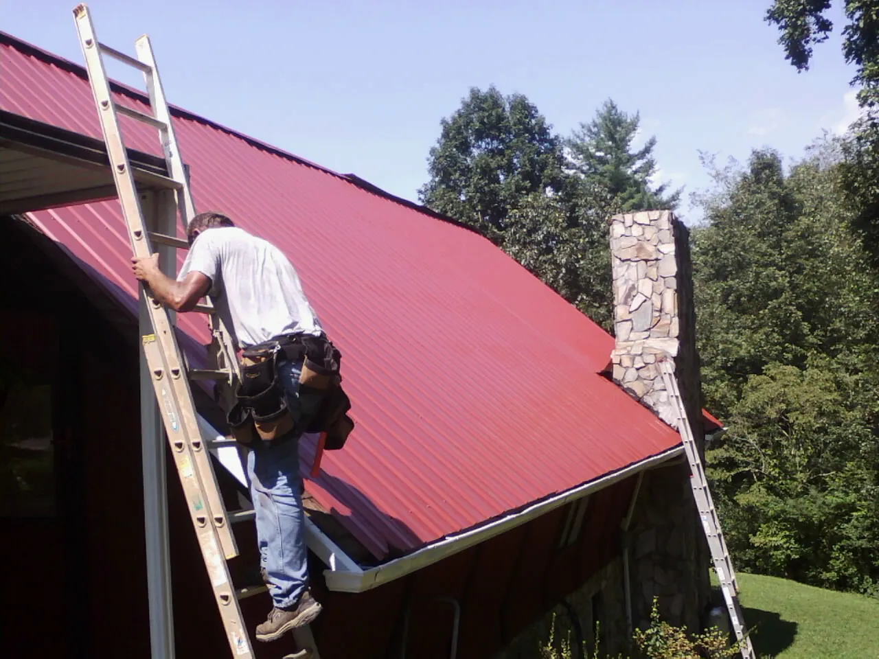 Roofer on a ladder beside a red metal roof and stone chimney — Soffit & Fascia Repair by Roofing Brothers in Youngsville